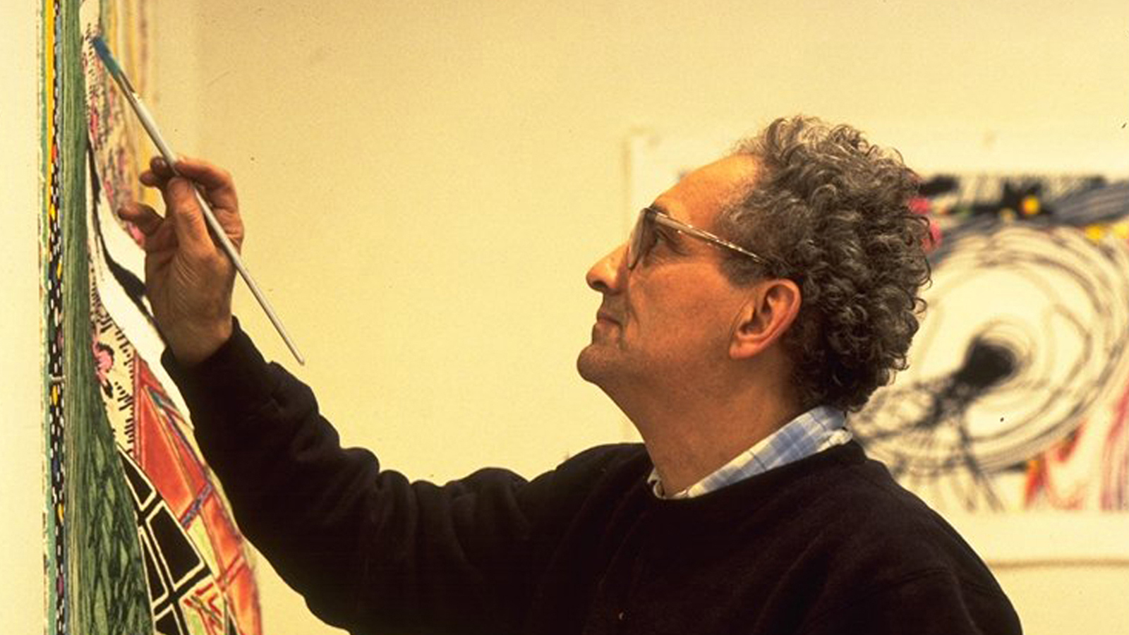 Color photo of the profile of a light skinned man with an arm upraised and hand holding a paintbrush to a canvas on the wall